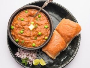 A plate of Pav Bhaji with buttery pav buns, garnished with peas and served with chopped onions and lime wedges.