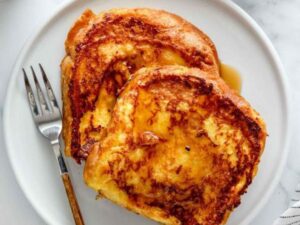 Two golden slices of french toast recipe drizzled with syrup on a white plate, next to a fork.